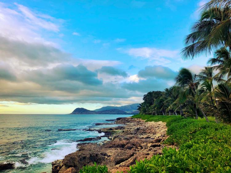 Blue skies and clouds near body of water alongside rocky terrain and green field and trees